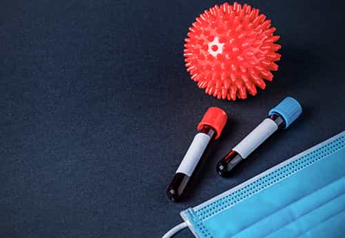 photograph of items on a desk including a red stress ball with spiky points like a covid molecule, vials with a red and a blue top, and a facemask
